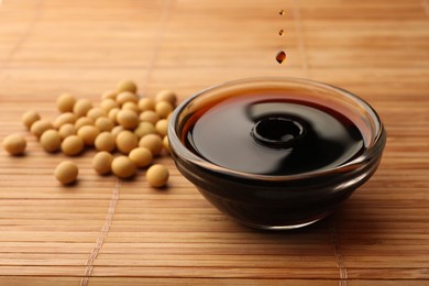 Photo of Soy sauce drops falling into bowl and soybeans on bamboo mat, closeup