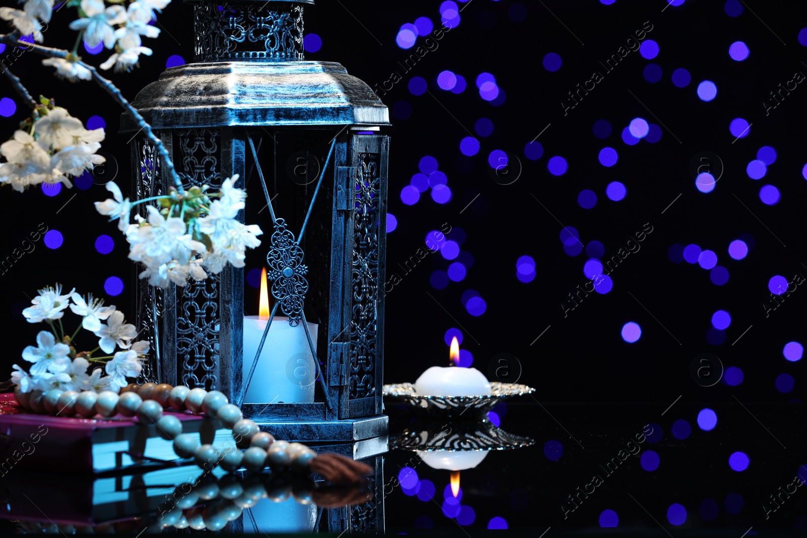 Photo of Arabic lantern, Quran, misbaha, burning candle and flowers on mirror surface against blurred lights at night. Space for text