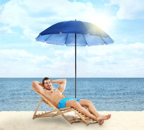 Young man on lounger under umbrella for sun protection at sandy beach 