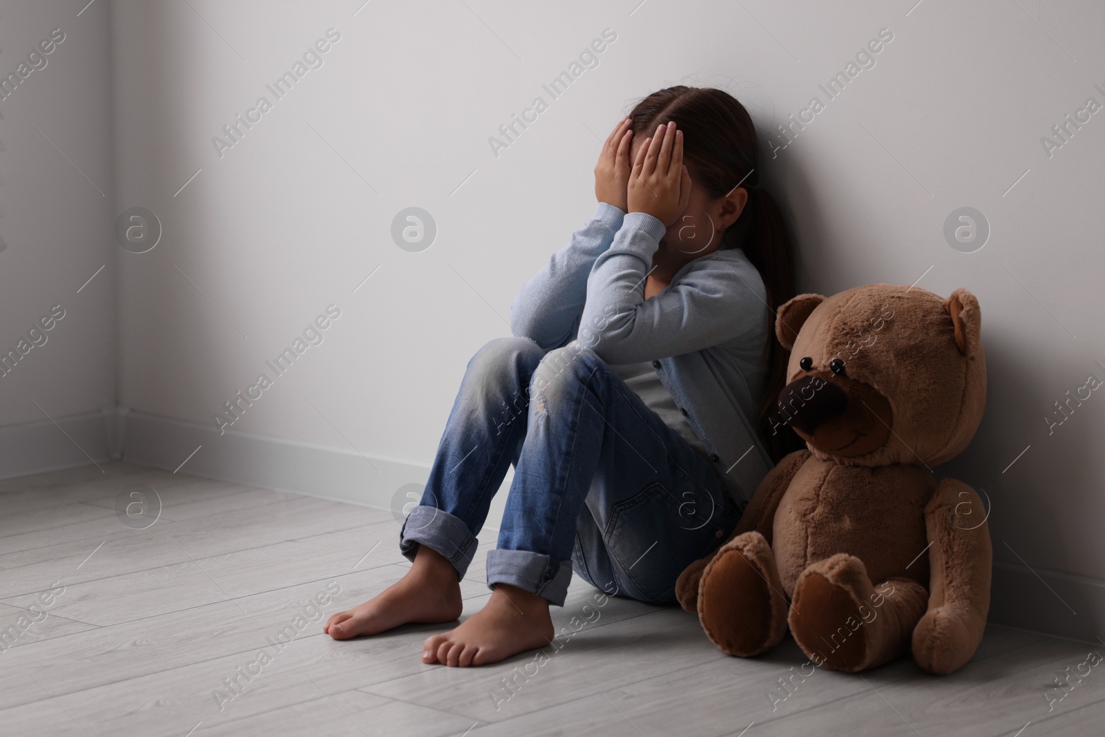 Photo of Child abuse. Upset little girl with teddy bear sitting on floor near light wall indoors, space for text