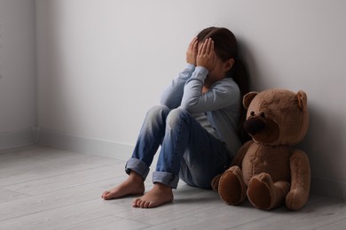 Photo of Child abuse. Upset little girl with teddy bear sitting on floor near light wall indoors, space for text