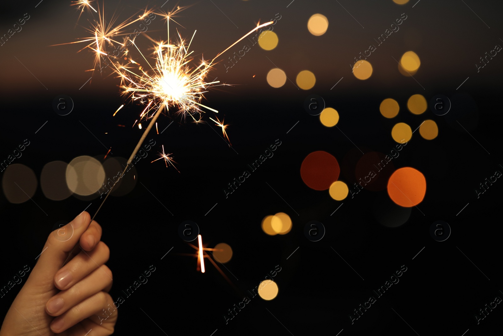 Photo of Woman holding bright sparkler against blurred lights, closeup. Space for text