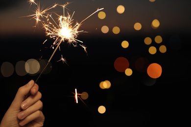 Woman holding bright sparkler against blurred lights, closeup. Space for text