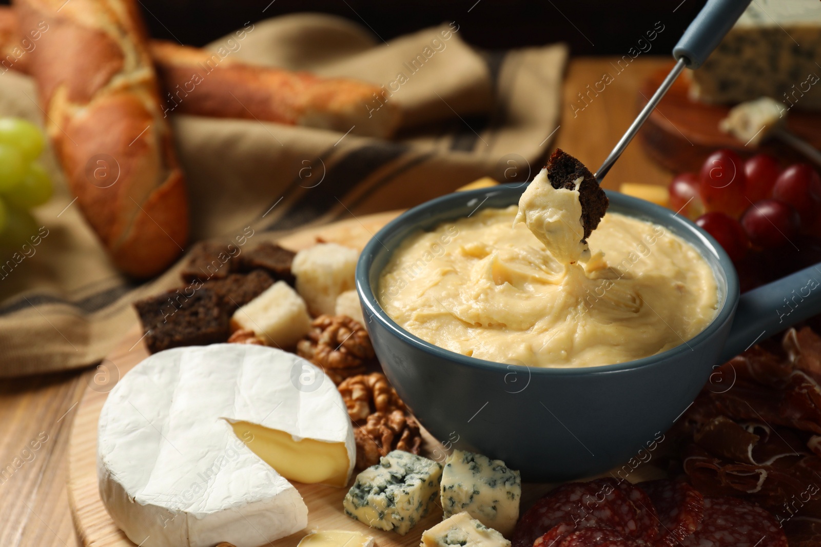 Photo of Dipping piece of bread into fondue pot with melted cheese at wooden table, closeup