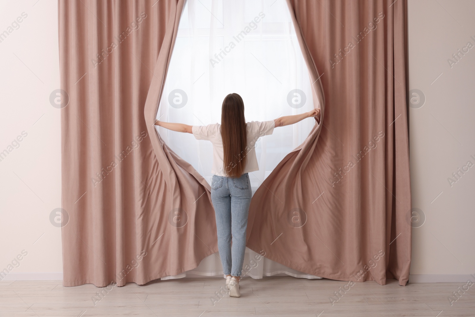Photo of Woman opening stylish curtains at home, back view