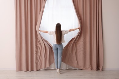 Woman opening stylish curtains at home, back view