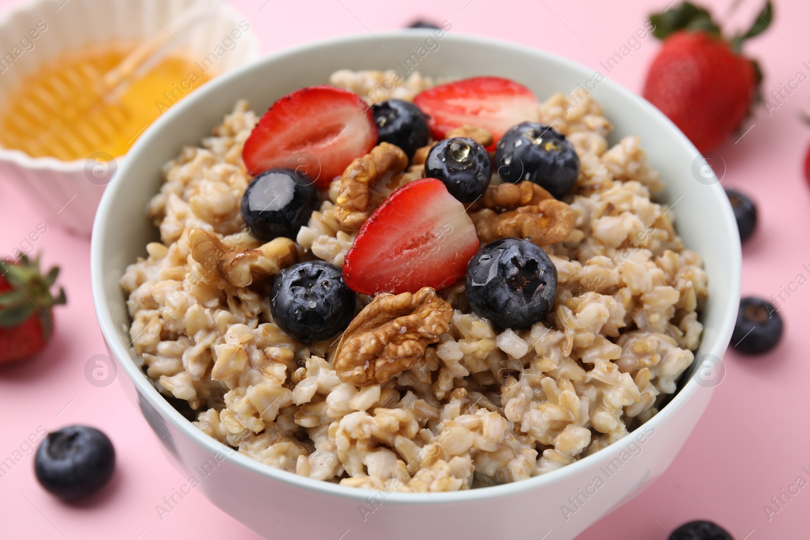 Photo of Tasty oatmeal with strawberries, blueberries and walnuts in bowl surrounded by fresh berries on pink background, closeup