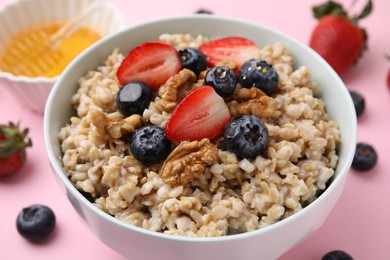 Photo of Tasty oatmeal with strawberries, blueberries and walnuts in bowl surrounded by fresh berries on pink background, closeup