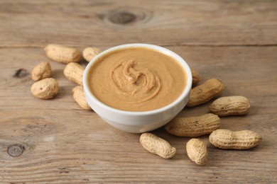 Photo of Delicious nut butter in bowl and peanuts on wooden table, closeup