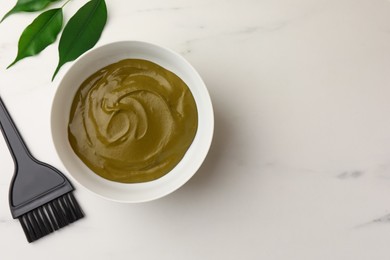 Bowl of henna cream, brush and green leaves on white marble table, flat lay with space for text. Natural hair coloring