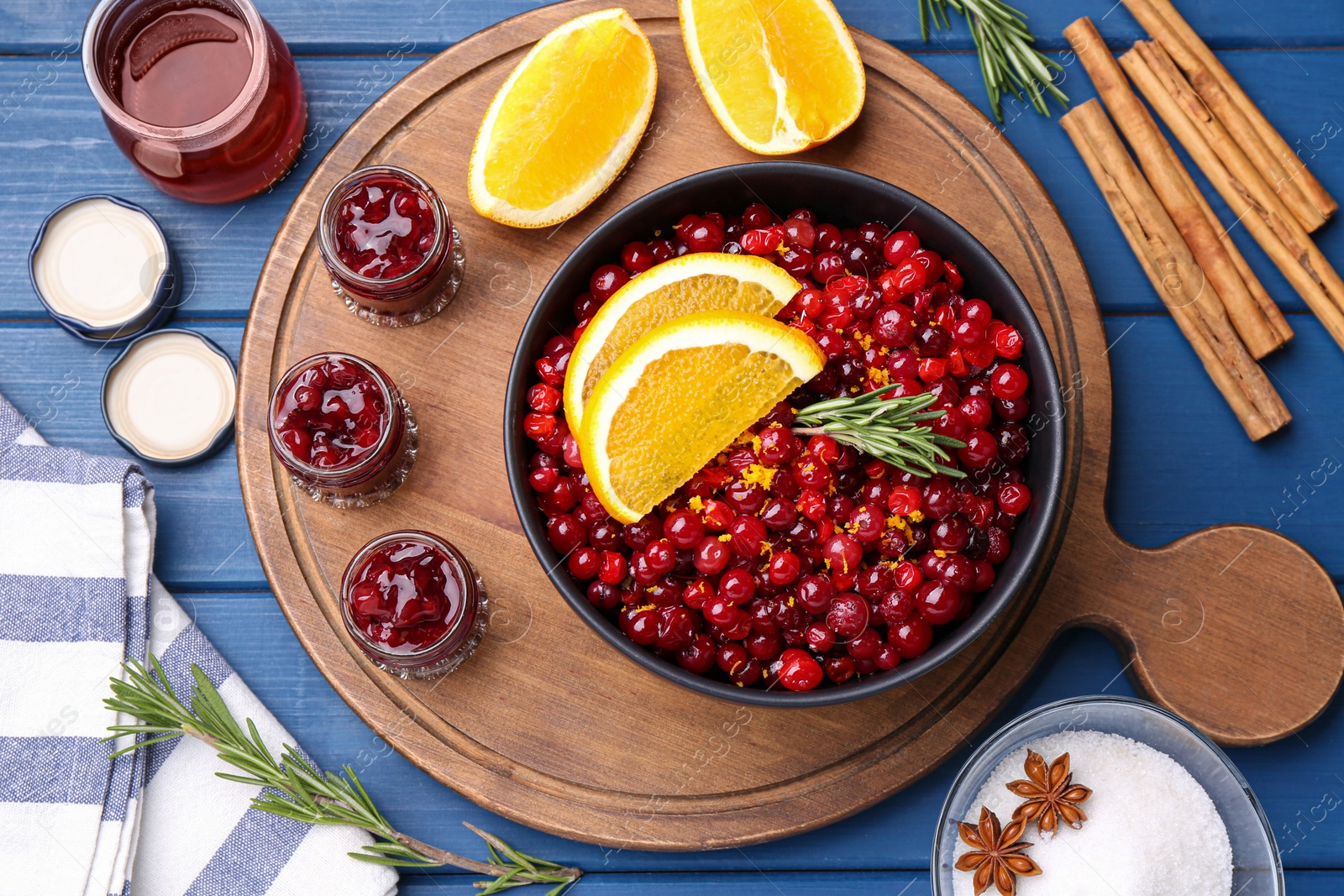 Photo of Cranberries in bowl, jars with sauce and ingredients on blue wooden table, flat lay