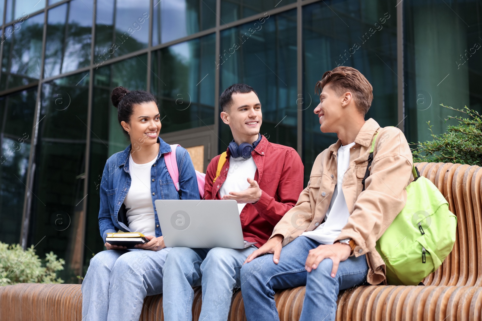 Photo of Happy young students studying with laptop on bench outdoors