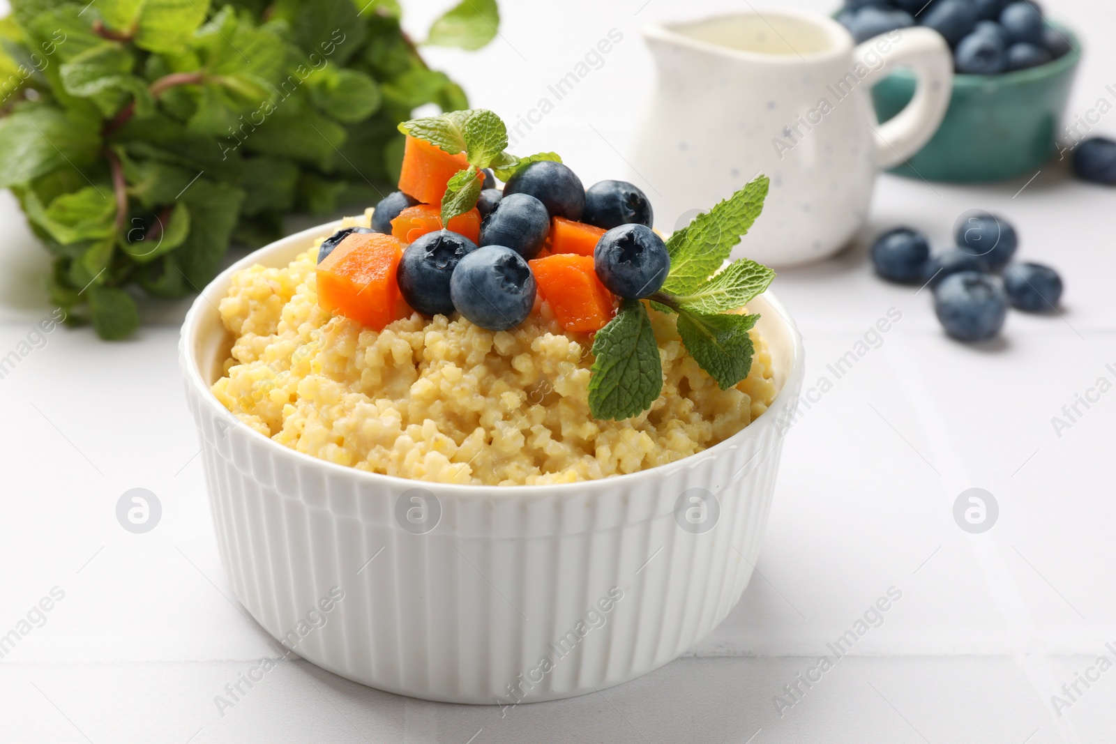 Photo of Tasty millet porridge with blueberries, pumpkin and mint in bowl on white tiled table, closeup