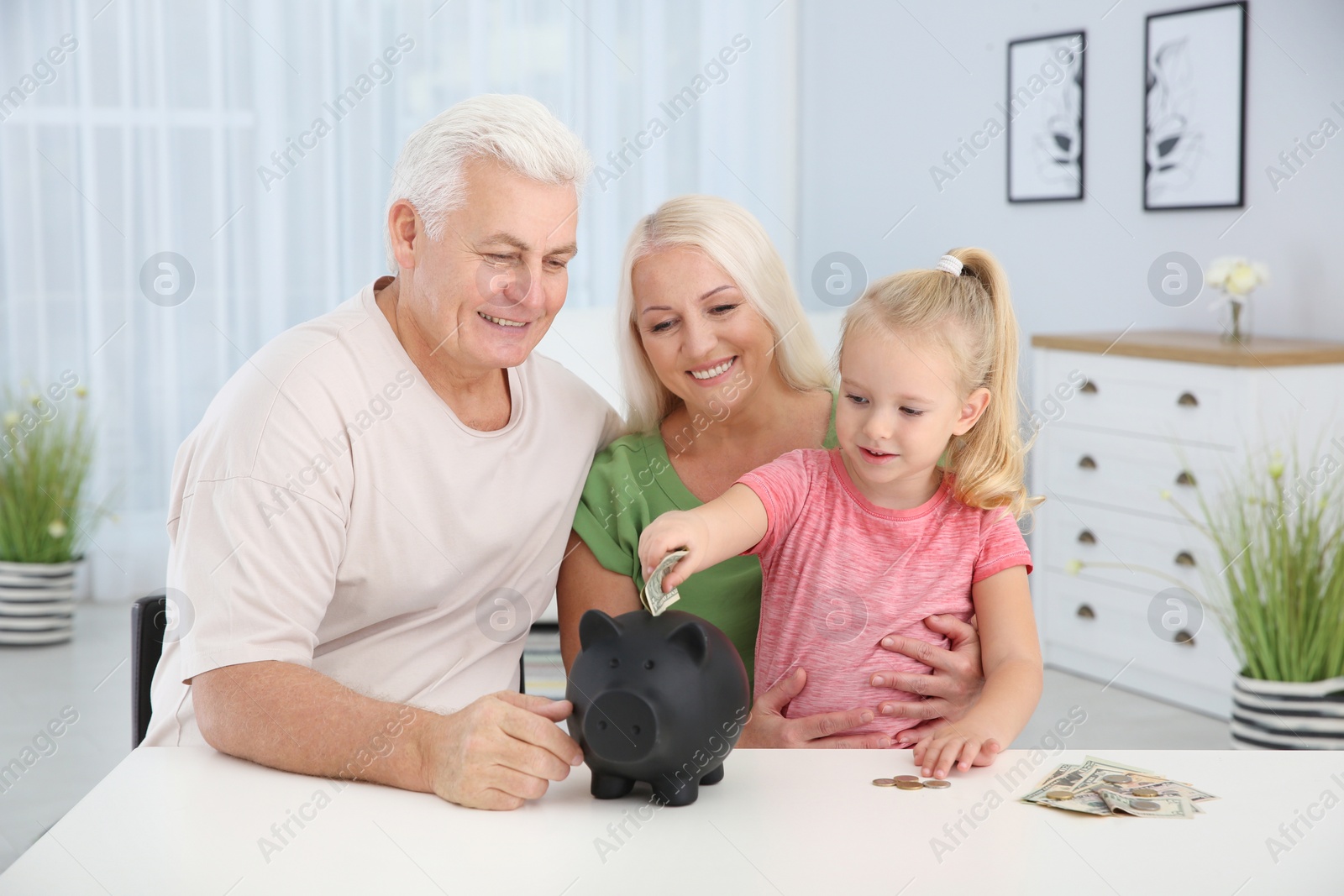 Photo of Little girl putting money into piggy bank and her grandparents at table