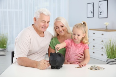 Photo of Little girl putting money into piggy bank and her grandparents at table