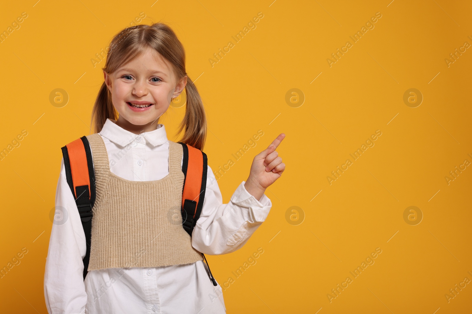 Photo of Happy schoolgirl with backpack pointing at something on orange background, space for text