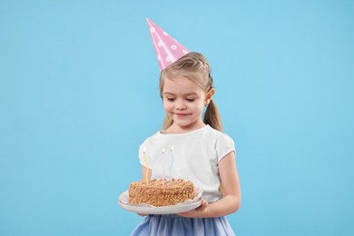 Birthday celebration. Cute little girl in party hat holding tasty cake with burning candles on light blue background