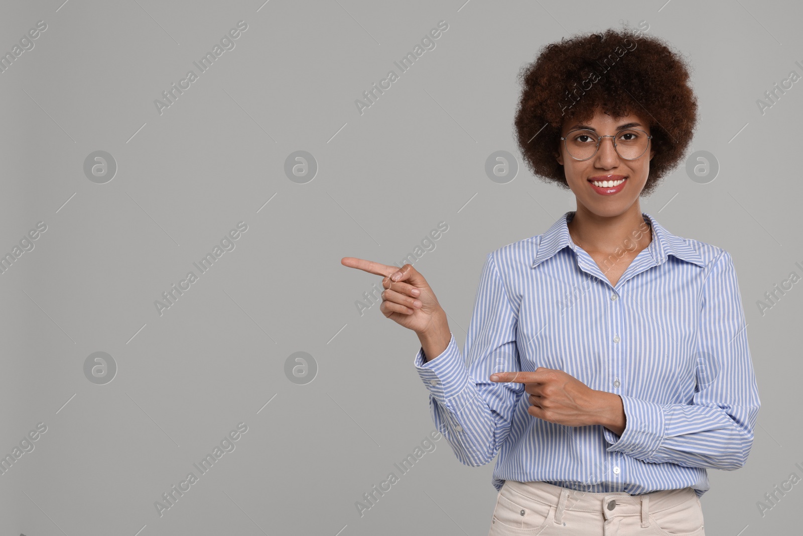 Photo of Happy young woman in eyeglasses pointing at something on grey background. Space for text