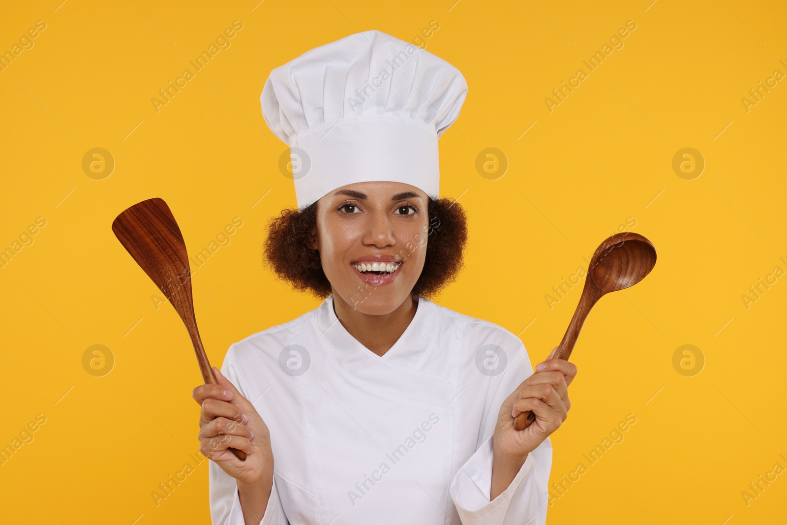 Photo of Happy female chef in uniform holding spoon and spatula on orange background