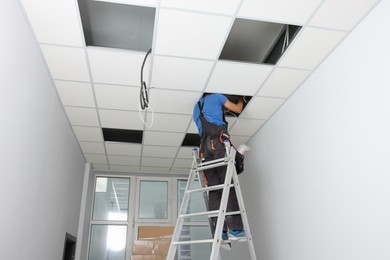Photo of Installing ceiling lighting. Electrician working on step ladder in room