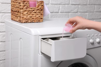 Photo of Woman pouring fabric softener from cap into washing machine near white brick wall, closeup