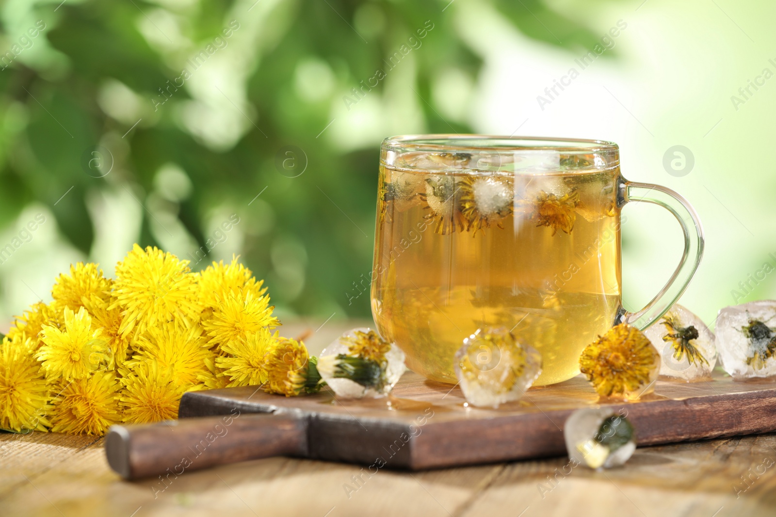 Photo of Delicious fresh tea, dandelion flowers and ice cubes on wooden table against blurred background