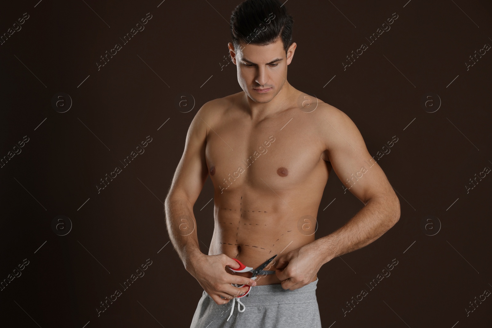 Photo of Fit man with scissors and marks on body against dark brown background. Weight loss surgery