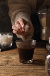 Photo of Brewing aromatic coffee in moka pot. Woman putting sugar cubes in glass with drink at wooden table, closeup