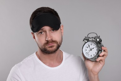 Photo of Sleepy man with alarm clock and mask on grey background. Insomnia problem