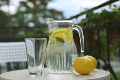 Jug with refreshing lemon water, glass and citrus fruits on light table outdoors