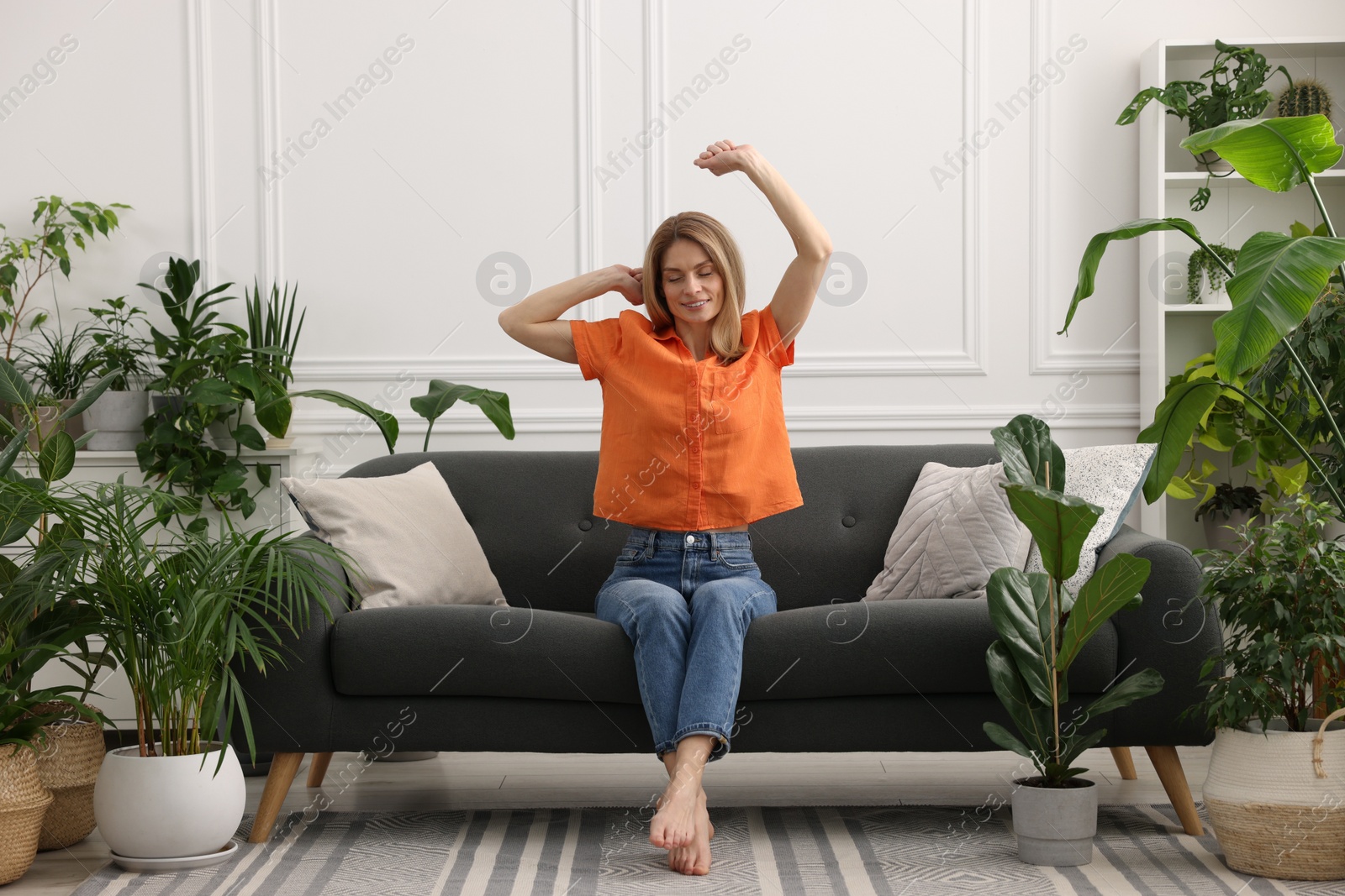 Photo of Woman stretching on sofa surrounded by beautiful potted houseplants at home
