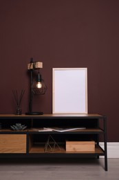 Photo of Empty frame and stylish lamp on wooden table near brown wall. Mockup for design