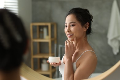 Photo of Happy woman applying face cream near mirror at home