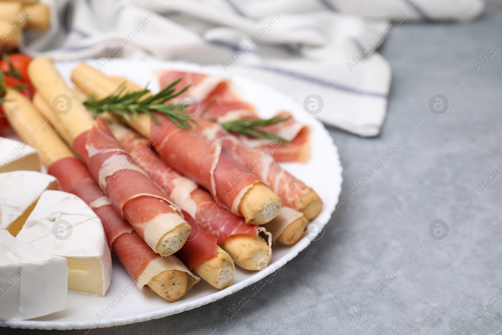 Photo of Plate of delicious grissini sticks with prosciutto, cheese and rosemary on light grey table, closeup. Space for text