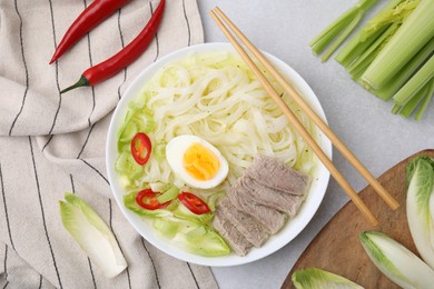 Photo of Bowl of delicious rice noodle soup with meat and egg near ingredients on light grey table, flat lay