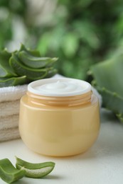 Jar with cream and cut aloe leaves on white table against blurred green background, closeup