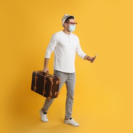 Male tourist n protective mask holding passport with ticket and suitcase on yellow background