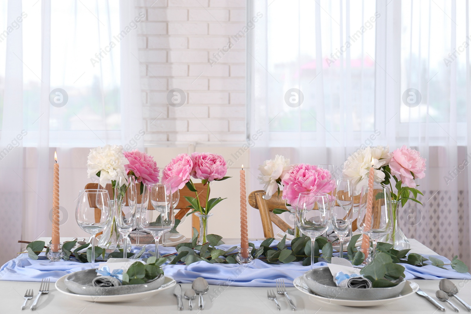 Photo of Beautiful table setting. Plates with greeting cards, napkins and branches near glasses, peonies, burning candles and cutlery on table in room