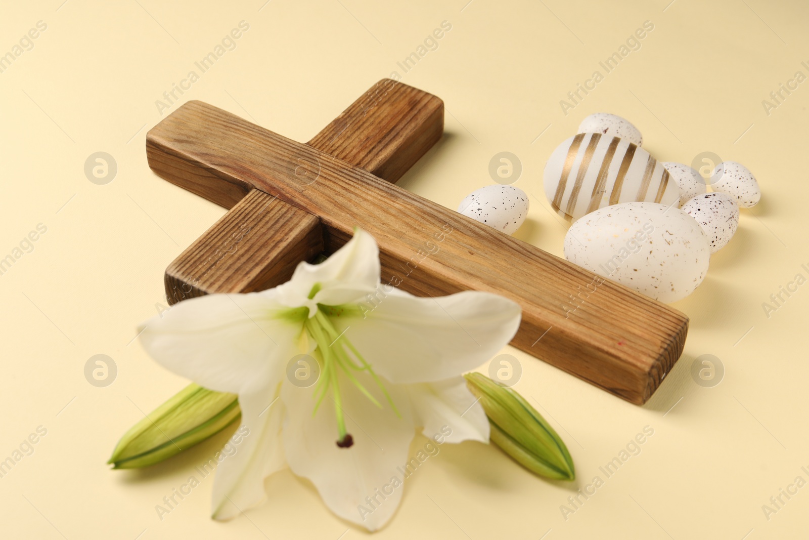 Photo of Wooden cross, painted Easter eggs and lily flowers on pale yellow background, closeup