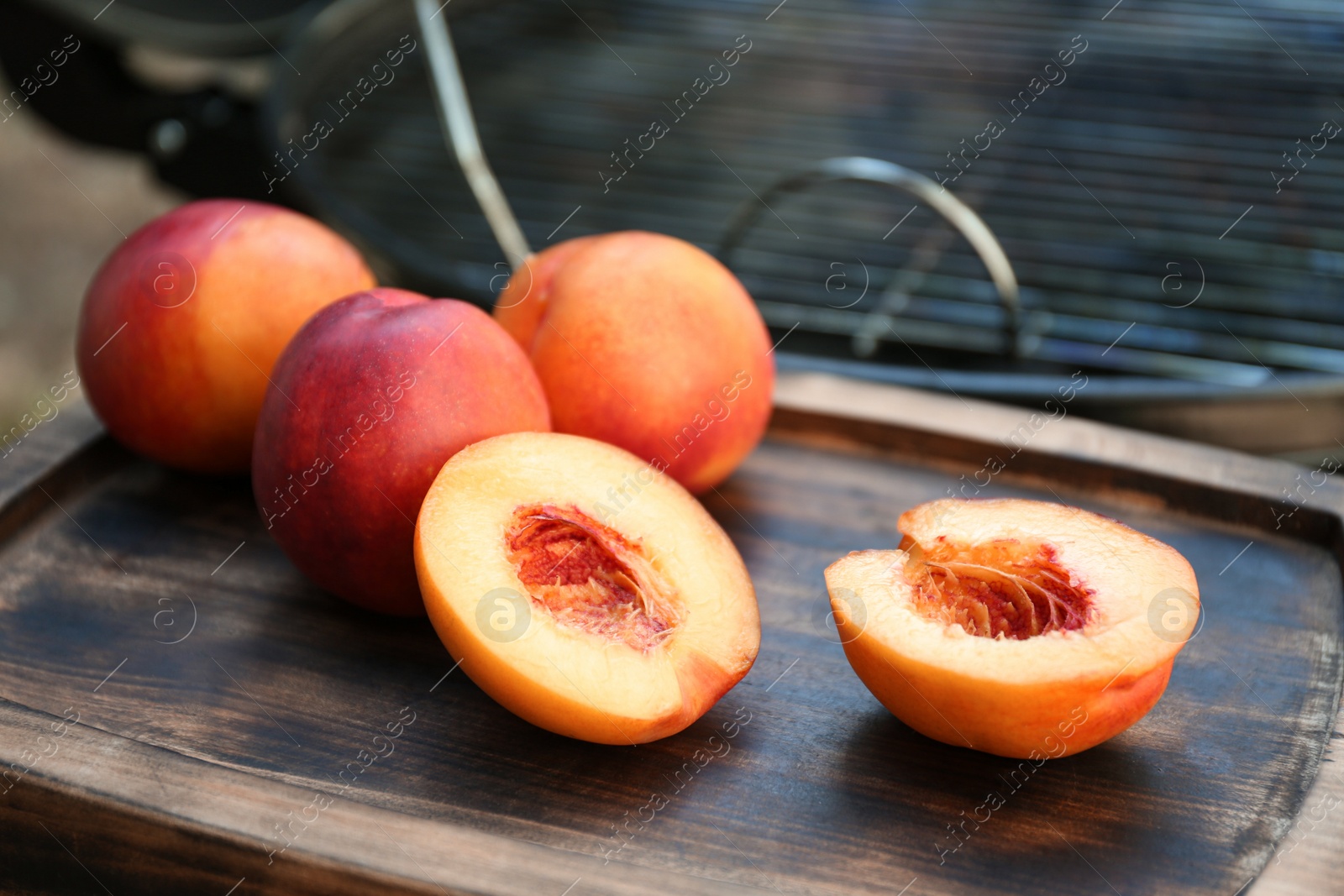 Photo of Fresh cut and whole peaches on wooden board near modern grill outdoors, closeup