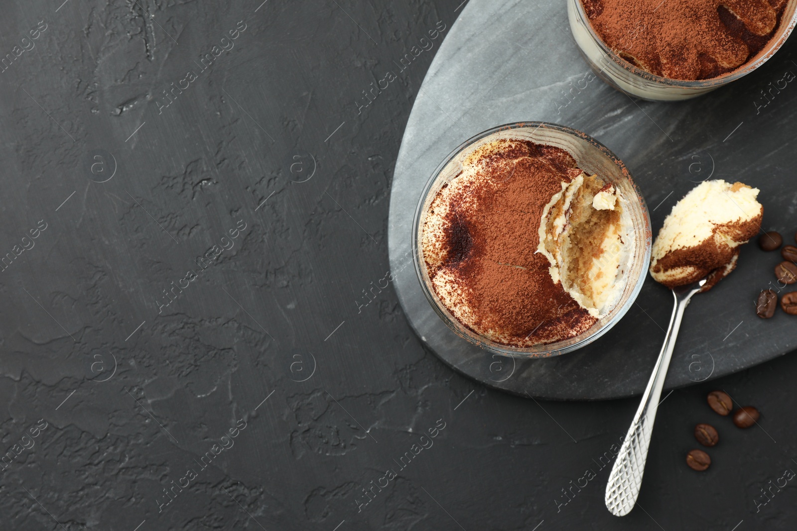 Photo of Delicious tiramisu in glasses, spoon and coffee beans on black table, top view. Space for text