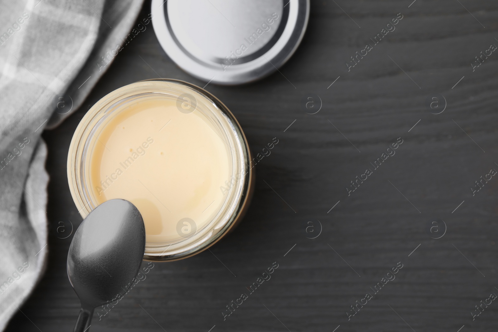 Photo of Open jar with condensed milk and spoon on dark grey wooden table, flat lay. Space for text