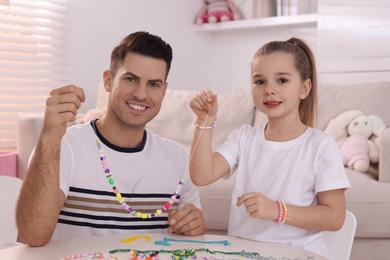 Photo of Happy father and his cute daughter with bright handmade beaded jewelry at table in room