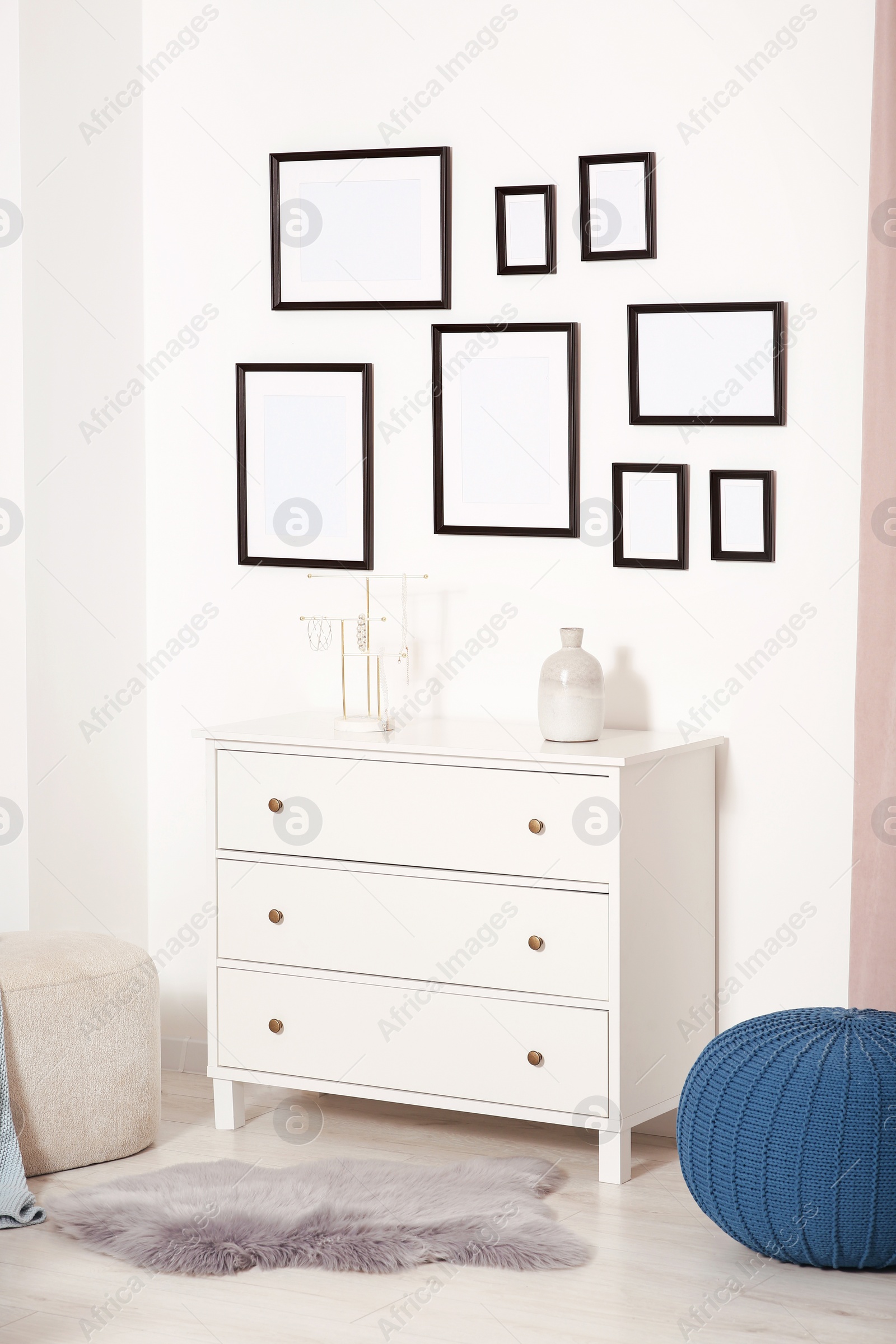 Photo of Empty frames hanging on white wall, chest of drawers and ottomans indoors