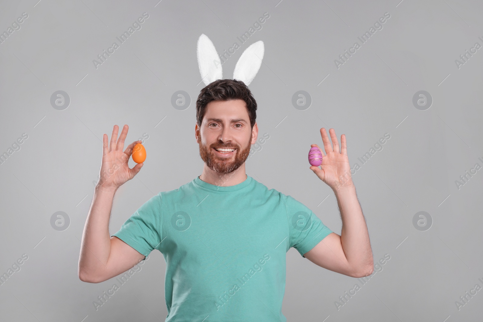 Photo of Portrait of happy man in cute bunny ears headband holding Easter eggs on light grey background