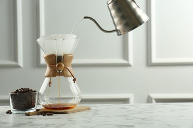 Photo of Pouring hot water from kettle into glass chemex coffeemaker on white marble table, space for text