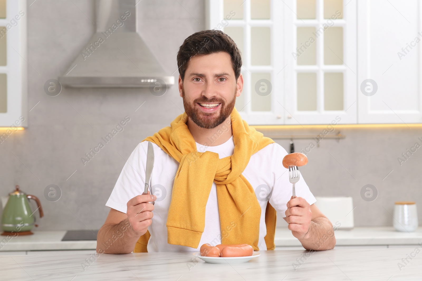 Photo of Happy man holding knife and fork with sausage at table in kitchen