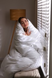 Beautiful young woman wrapped with soft blanket near window at home