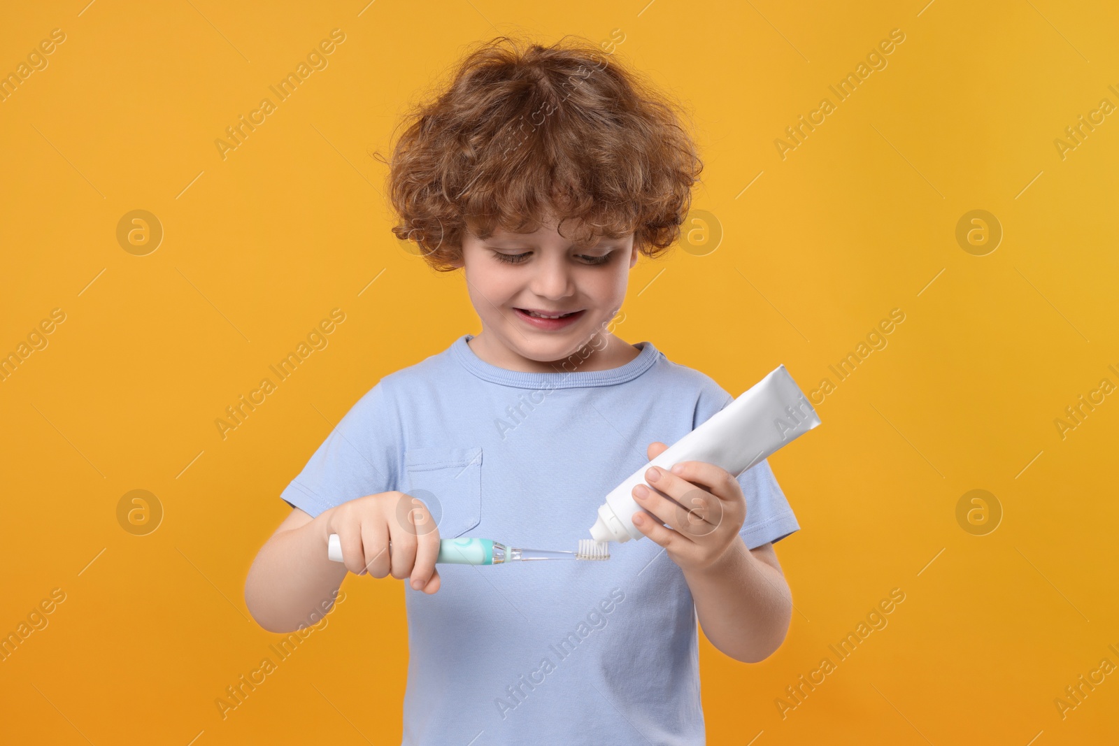 Photo of Cute little boy squeezing toothpaste from tube onto electric toothbrush on yellow background