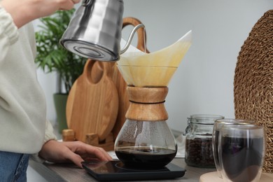 Photo of Woman pouring hot water into glass chemex coffeemaker with paper filter and coffee at countertop in kitchen, closeup